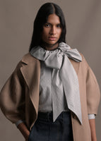 A zoomed in image of model wearing a long camel regency coat with short balloon sleeves over a striped blouse with a neck tie.