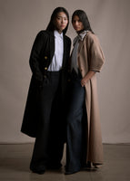 Two models: The model on the right is wearing a black coat with black pants and white top, the model on the right wearing a long camel regency coat with short balloon sleeves over a striped blouse with neck tie and pants.