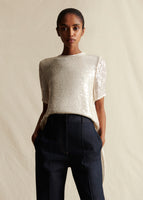 Model facing forwards wearing an ivory sequin short sleeve top with dark denim jeans.