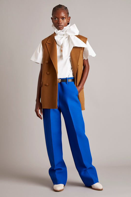 Fall 2021 main image, a model wearing blue trousers, a white shirt with a big neck bow and a brown sleeveless blazer 