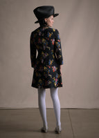 A back-facing image of a model wearing a short black multicolor flower print long sleeve dress with a black hat.