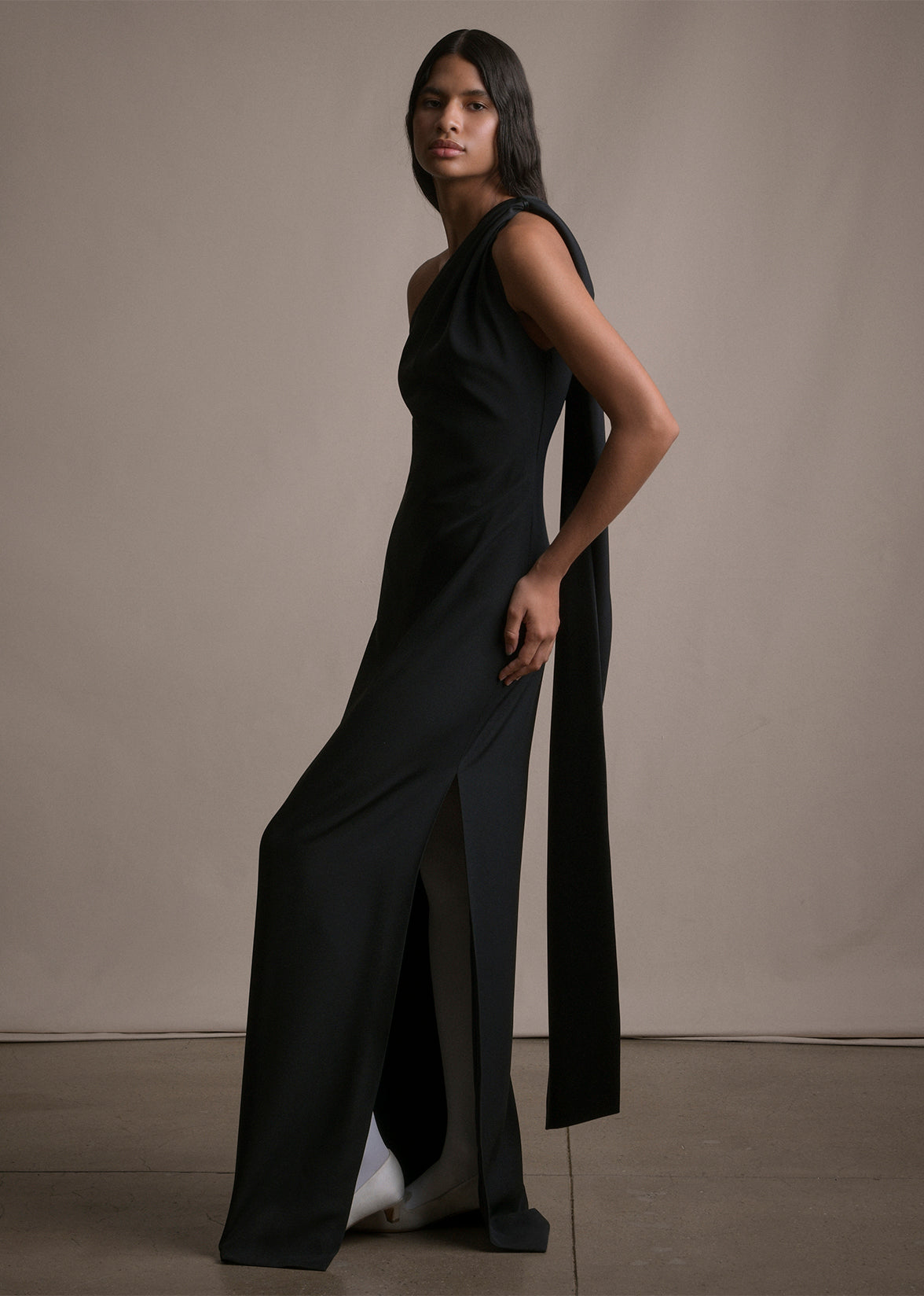 Side angle image of model wearing floor length black satin one shoulder gown with wrap ribbon detail and back slit.