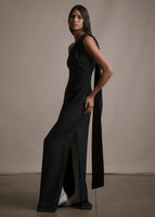 Side angle image of model wearing floor length black satin one shoulder gown with wrap ribbon detail and back slit.