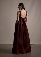 A back-facing image of a model wearing a floor length fit and flare merlot gown with thin straps and gathered bow-like on the front torso.