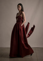 Side angle image of model wearing a floor length fit and flare merlot gown with thin straps and gathered bow-like on front torso