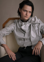 A zoomed in image of model wearing a long sleeve blouse with high neckline in stripe shirting with a tied bow at the front neck.