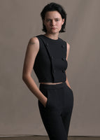 Model in a cropped sleeveless black gilet top with six black buttons on the front paired with black pants. 