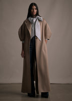 Model wearing a long camel regency coat with short balloon sleeves over a bow tied striped blouse.