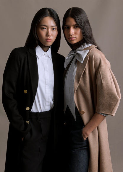 An image of two models, one wearing a black double breasted coat over white collared shirt. The other is wearing a camel colored coat over a striped shirt by Adam Lippes