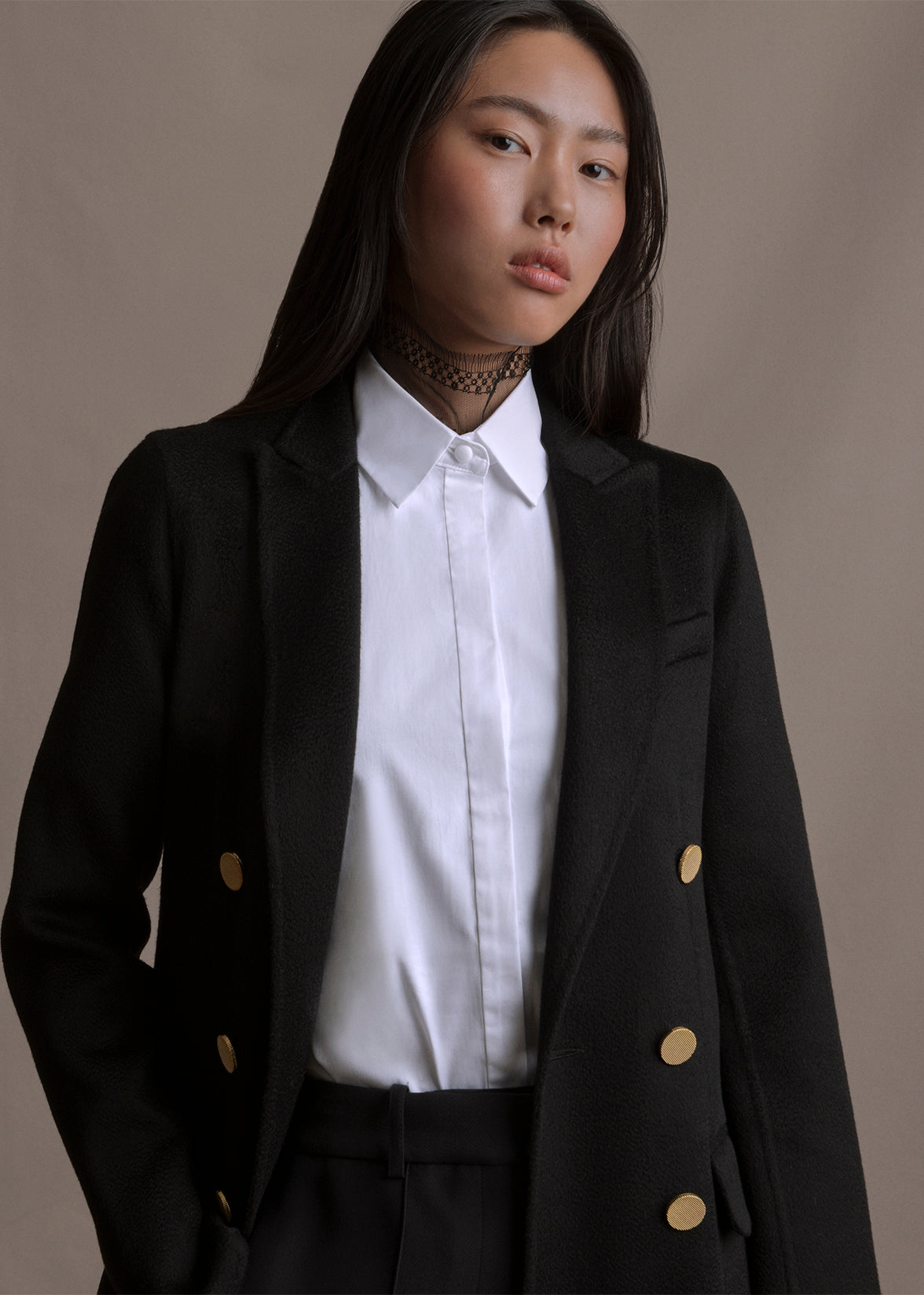 A close-up image from the waist up of a model wearing a black double breasted coat over white collared shirt by Adam Lippes