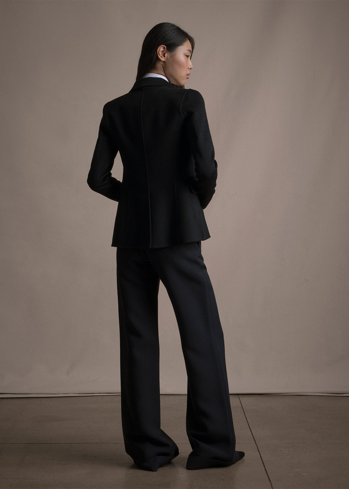 Image of a model standing backwards wearing a black blazer with black trousers by Adam Lippes