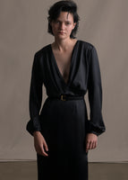 A zoomed in image of model wearing a black satin dress with a v-neckline and a black belt with a gold buckle.