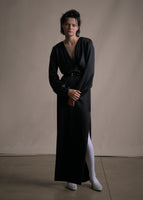 Model wearing a long sleeve floor length black satin dress with a v-neckline and a black belt with a gold buckle. 