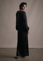 A back-facing image of a model wearing a long sleeve floor length satin dress tied with a gold chain.