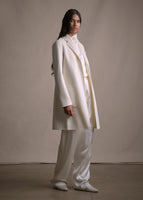 A model standing sideways wearing an in ivory long sleeved coat over ivory pants.