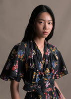 A zoomed in image of model wearing a mid length black short sleeve asymmetrical dress with a tie front and multicolor printed flowers.