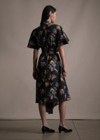 A back-facing image of model wearing a mid length black short sleeve asymmetrical dress with a tie front and multicolor printed flowers.