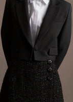 A zoomed in image of model wearing short black metallic wrap skirt with three black buttons paired with cropped blazer and white top.