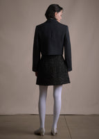 A back-facing image of model wearing short black metallic wrap skirt with three black buttons paired with cropped blazer.