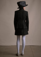 A back-facing image of a model wearing a mid length black metallic jacket with a matching skirt and a black hat.