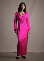 Model wearing a long sleeve floor length pink satin dress with a v-neckline and a pink belt with a gold buckle.