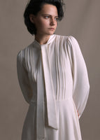 A zoomed in image of a white long sleeved silk dress with a self fabric bow at the front neck.