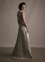 A side facing image of model wearing a  floor length champagne V-neck sleeveless gown in flower detail.