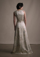 A back-facing image of a model wearing a  floor length champagne V-neck sleeveless gown in flower detail.