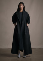 A model wearing a long black regency coat with short balloon sleeves and a front waist tie.