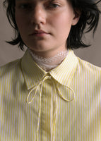 A zoomed in image of a model wearing white and citrine long sleeve shirt with collar and thin bow in stripe shirting.