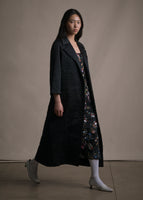 A side angle image of model wearing long black quilted opera coat with two pockets.