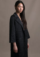 A side angle image of model wearing long black quilted opera coat with two pockets.