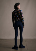 Model facing backwards wearing black floral printed long sleeved shirt tucked into navy blue flare pants.