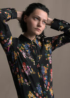 A zoomed in image of model wearing a black long sleeve shirt with collar, thin bow, and multicolor printed flowers.
