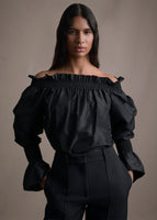 Cropped image of a model wearing a black silk off the shoulder top tucked into black pants.