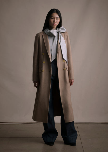 An image of a model facing forward wearing a camel double breasted long coat over a striped top with a bow and with blue jean pants by Adam Lippes