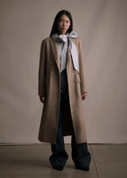 An image of a model facing forward wearing a camel double breasted long coat over a striped top with a bow and with blue jean pants.