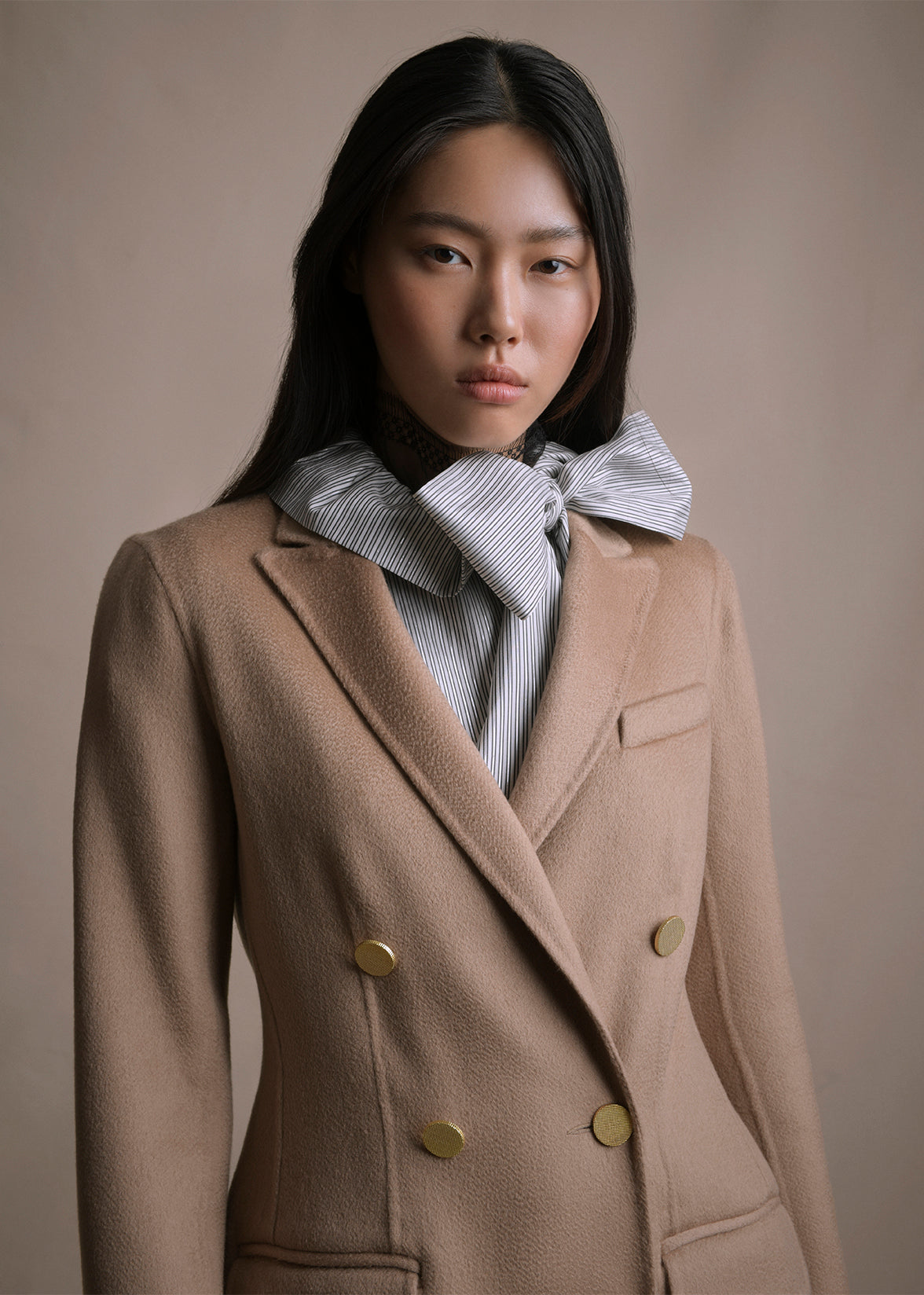 Close up image from waist up of a model wearing a camel double breasted jacket over a striped top by Adam Lippes