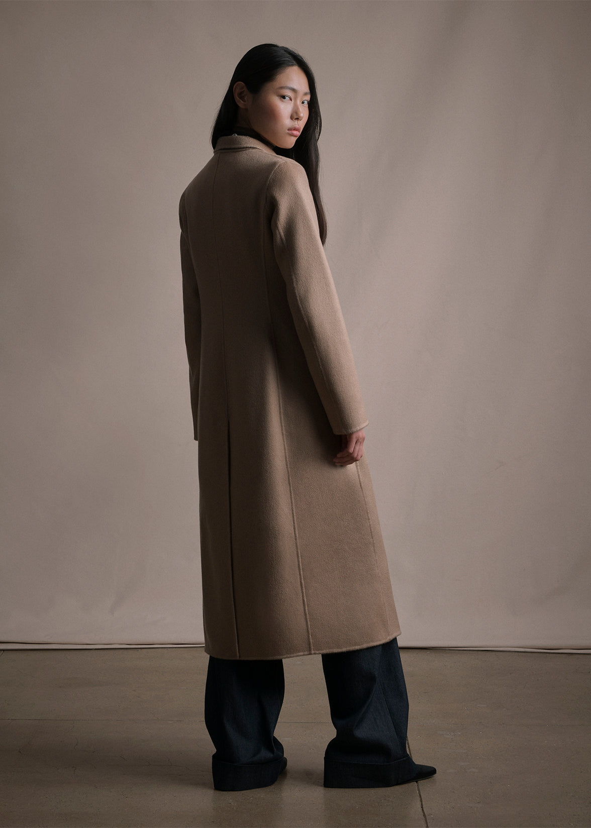 Image of a model standing backwards wearing a camel long coat over blue denim jeans by Adam Lippes