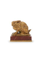 A bronze model of a crouching rabbit with its ears down on a red stone base.