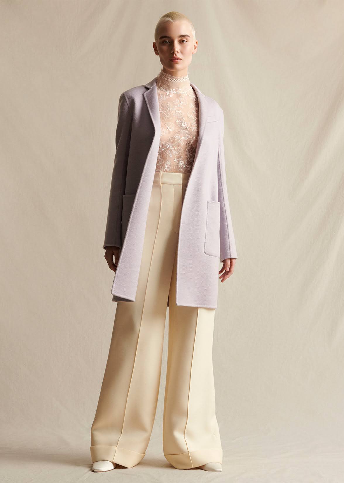 A model wearing a lilac mid-length coat, layered over an ivory lace turtleneck.  Styled with ivory wide leg pants.