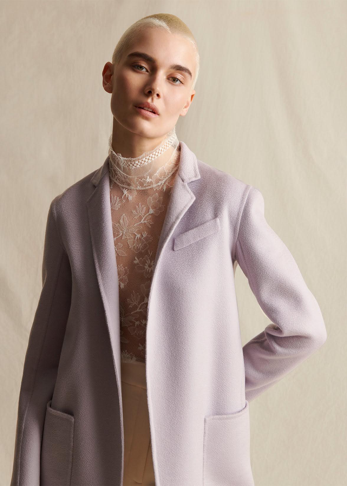 A model wearing a lilac mid-length coat, layered over an ivory lace turtleneck.  Styled with ivory wide leg pants.