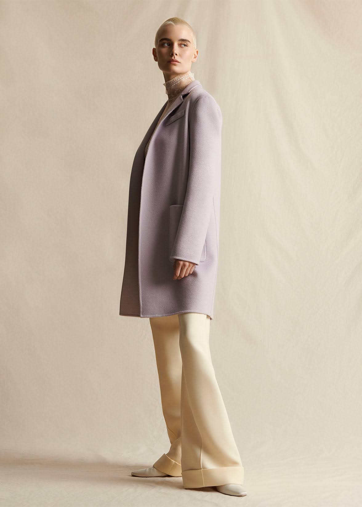 A model wearing a lilac mid-length coat, layered over an ivory lace turtleneck.  Styled with ivory wide leg pants.