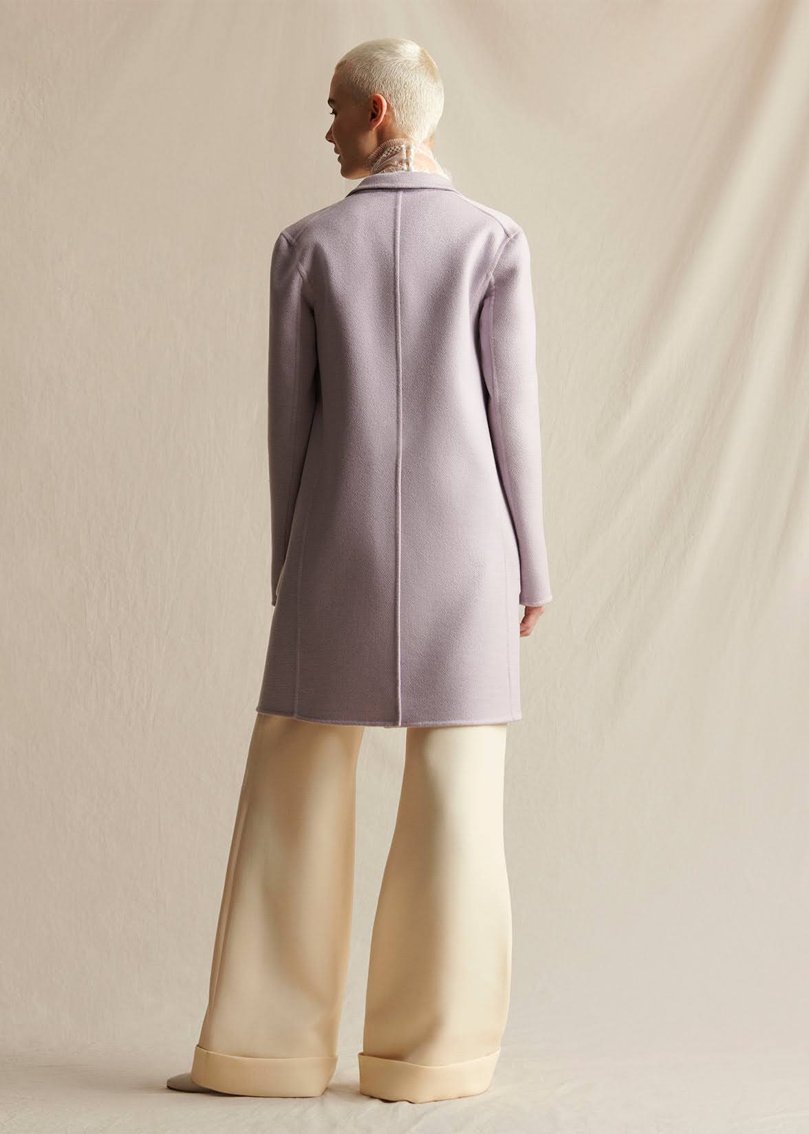 A model wearing a lilac mid-length coat, layered over an ivory lace turtleneck.  Styled with ivory wide leg pants.