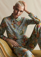 A model sitting on a yellow bench wearing a pale blue floral silk long sleeved top and matching pants. 