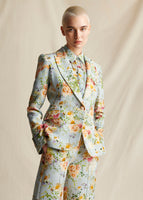 An image of a model wearing a pale blue floral printed blazer, with matching pants and collared shirt.