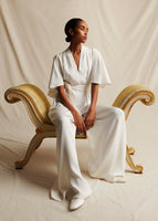 An image of a model wearing an ivory silk V-neck jumpsuit while sitting on an antique bench.