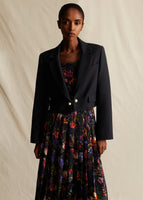 A model stands wearing a black cropped blazer, over a colorful printed pleated dress.