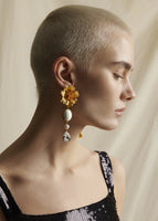 Close up image of a model wearing an earring with a large gold flower at the top, then a white stone, then a crystal.