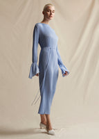 A side view of a model wearing a light blue pleated midi length dress with flounce cuff and self tie at the waist.
